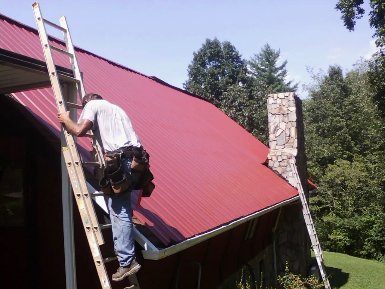 Roofer on a ladder beside a red metal roof and stone chimney — Soffit & Fascia Repair by Roofing Brothers in Curlew